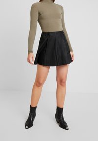 Jupe plissée en cuir noir associée à un col roulé ajusté en olive côtelé. Modèle portant des bottines noires avec des accents en argent.