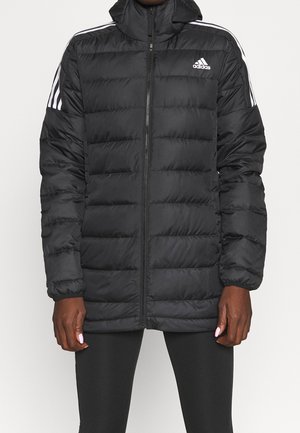 Personne portant une veste matelassée noire Adidas avec des bandes blanches sur les épaules et les manches, fermée par une fermeture éclair, debout devant un fond uni.