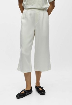 Osoba nosząca luźne, białe spodnie o długości do połowy łydki oraz czarne, płaskie buty w stylu Mary Jane, stojąca na tle białego tła.