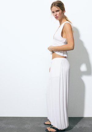 Mladá žena v voľnom bielom tielku bez rukávov a širokých bielych nohaviciach stojaca bokom pri bielej stene, obutá v čiernych sandáloch.