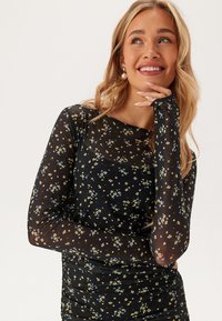 Sort mesh-langærmet top med et blomstermønster i gult og blåt. Har en rund halsudskæring og rynket kant for en tætsiddende silhuet.