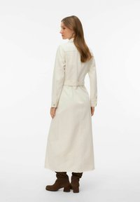 Femme aux longs cheveux bruns portant un manteau long crème et des bottines marron, debout de dos sur un fond blanc.