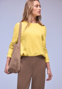Femme portant un haut jaune à manches longues et un pantalon marron, avec un sac à bandoulière beige, regardant vers la droite sur un fond bleu clair.