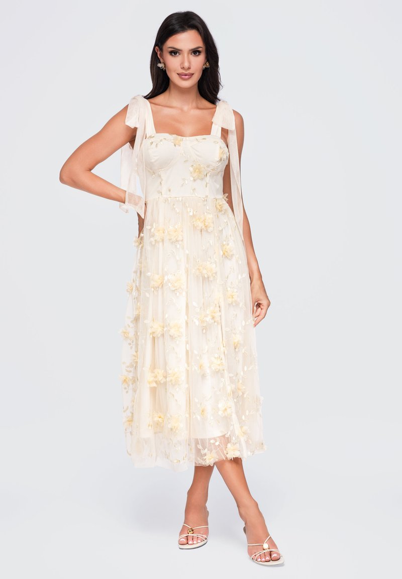 Femme portant une robe crème sans manches avec des appliques florales et des bretelles nouées, debout, une main sur la hanche, chaussant des sandales.
