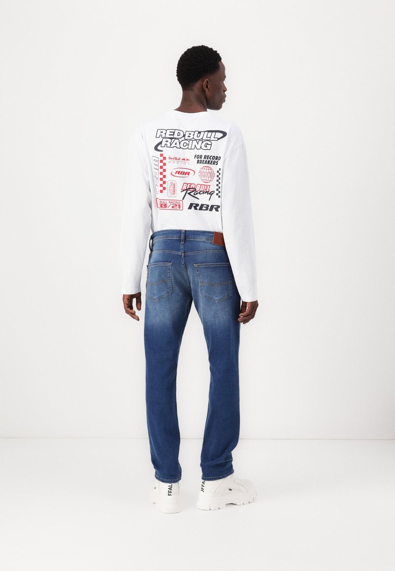 Weißes Langarmhemd mit "Red Bull Racing" Grafik auf dem Rücken, kombiniert mit blauen Jeans und weißen Turnschuhen, von hinten gesehen.