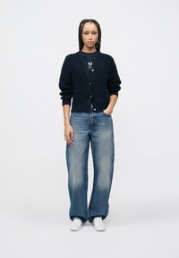 Πλεκτό μπλε σκούρο ζακέτα με κούμπωμα κουμπιών και μπροστινές τσέπες, συνδυασμένο με άνετο ανοιχτό μπλε τζιν και άσπρα αθλητικά παπούτσια.