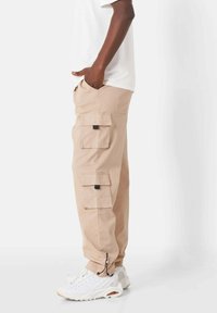 Beige cargobroek gemaakt van katoen, met meerdere zijzakken, zwarte hardware en verstelbare boorden. Gelijkend met witte sneakers.