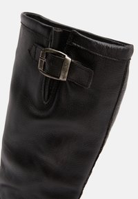 Botte noire en cuir allant jusqu'au genou avec une texture lisse, dotée d'un accent en boucle près du haut et de détails de couture minimaux.