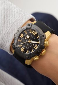Orologio cronografo nero e oro con grandi indicatori numerici, dettagli in acciaio inossidabile e cinturino in gomma, mostrato su un polso.
