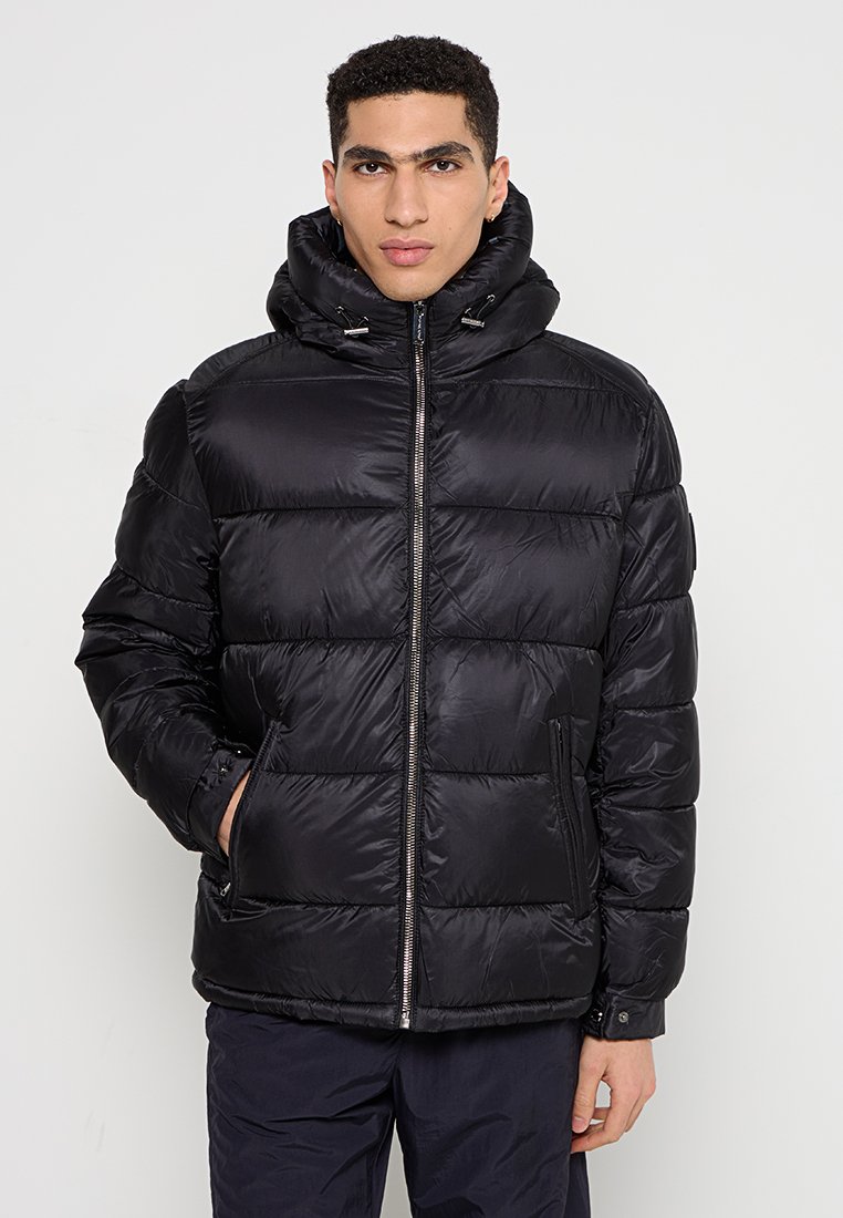 Uomo con capelli ricci e corti che indossa un giubbotto imbottito trapuntato nero con cappuccio, in piedi con una mano in tasca su uno sfondo neutro.