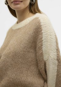 Pull en tricot beige avec un col crème et un accent crème clair sur une manche. Tissu texturé avec des détails de couture visibles.