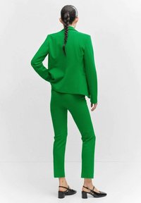 Zielony garnitur z dopasowanej tkaniny, składający się z dopasowanej marynarki i wąskich spodni, uzupełniony czarnymi szpilkami z kostką.