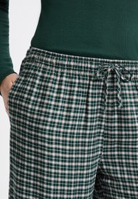 Persoon in groen-witte geruite broek met een trekkoord in de taille en een donkergroene lange mouwtop, met hand in de zak.