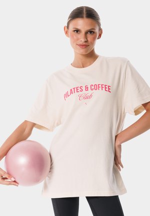 Kobieta z włosami związanymi w kok, ubrana w białą koszulkę z napisem „Pilates & Coffee Club”, trzyma różową piłkę do ćwiczeń, jedną ręką opartą na biodrze.