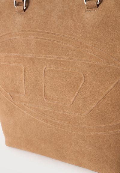 Gros plan d’un sac à main en daim couleur fauve avec des motifs géométriques abstraits en relief et des poignées cousues avec des anneaux métalliques.
