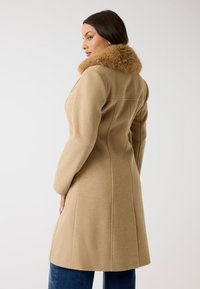 Cappotto di lana beige con colletto in pelliccia sintetica, design sartoriale e maniche lunghe. Tessuto strutturato con cuciture laterali e taglio a lunghezza media.