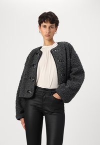 Veste en teddy noir texturé avec un col arrondi, des fermetures à l'avant et des manches volumineuses, associée à un pantalon noir ajusté et un haut crème.
