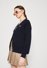 Ναυτικό πουλόβερ με V-λαιμόκοψη και κούμπωμα, με ένα μικρό πράσινο λογότυπο. Συνδυάζεται με μια πλισέ χρυσή φούστα. Χοντρές γυαλιά ηλίου.