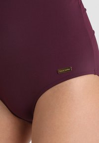 Bas de maillot de bain bordeaux en tissu lisse, avec une petite étiquette logo dorée sur le côté, design ajusté et bords sans coutures.
