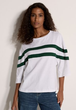 Mujer con cabello oscuro y rizado, vistiendo una camiseta blanca extragrande con dos franjas horizontales verdes oscuras y jeans azules, de pie contra una pared lisa.