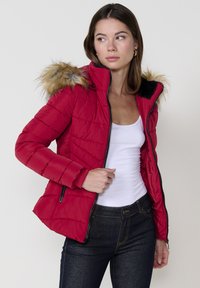 Raudona pūkinė striukė su užtrauktuku priekyje, juodais akcentais ir dirbtinio kailio kapišonu. Apsaugota šoninėmis kišenėmis su užtrauktuku ir išsiuvinėtu tekstūru.