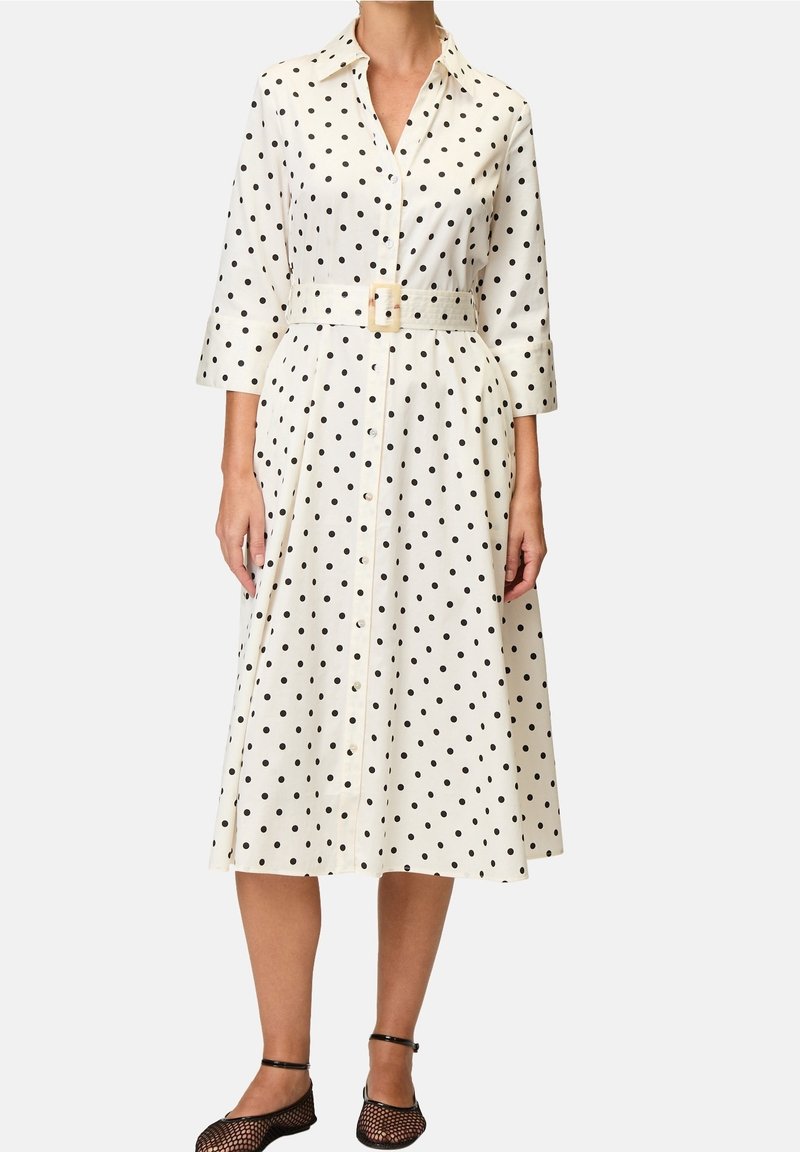 Robe midi blanche à pois noirs, devant boutonné, col chemise, manches 3/4, ceinture avec boucle, portée avec des chaussures à bride à la cheville en résille noire.
