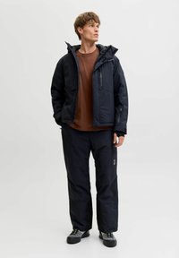 Veste noire isolée avec capuche et poches zippées, associée à un pantalon noir ample assorti. Chemise marron en dessous.