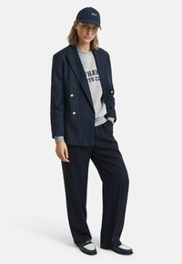 Blazer doppiopetto a righe navy sopra una felpa grigia chiara, abbinato a pantaloni scuri e mocassini navy con un leggero accento.