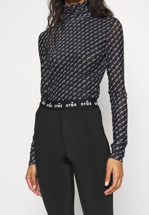 Musta istuva poolkauluspaita, jossa valkoinen vino "crās"-tekstikuvio ja siihen sopiva vyötärönauha, yhdistetty korkeavyötäröisiin mustiin housuihin.