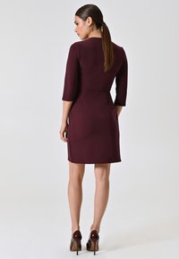 Bordeaux-farbenes, enges Kleid mit 3/4-Ärmeln, glatter Textur und strukturierter Silhouette. Getragen mit glänzenden High Heels, die eine schicke Rückenansicht zeigen.