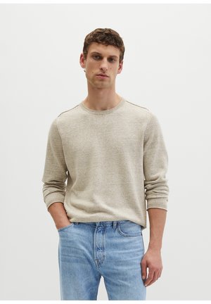 Młody mężczyzna w beżowym dzianinowym swetrze i jasnoniebieskich dżinsach stoi z jedną ręką w kieszeni na jednolitym tle.