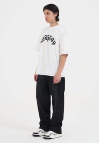 Hvid oversized t-shirt med sort skriftskrift, parret med mørke brede bukser og hvide sneakers med sorte og grå accenter.