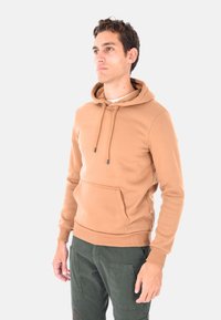Uomo con una felpa con cappuccio beige e pantaloni cargo verdi, in piedi e che guarda di lato su uno sfondo bianco uniforme.