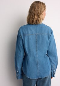 Denim blouse in lichtblauw, met lange mouwen, een naad op het achterpand en knoop manchetten. Gladde textuur met subtiele stiksel details.
