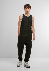 Schwarzes ärmelloses Tanktop mit weißem Saum, kombiniert mit schwarzen Sweatpants mit elastischen Bündchen und Seitentaschen, getragen mit grauen Turnschuhen.