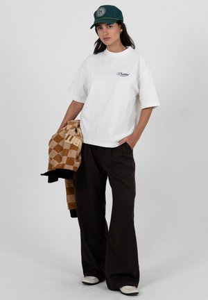 Vrouw met een wit T-shirt, zwarte wijde broek, witte schoenen en een groene pet, die in één hand een bruin geruit jasje vasthoudt.