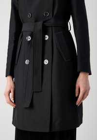 Manteau noir stratifié avec un design à double boutonnage, boutons en argent, ceinture à la taille et poches latérales. Tissu lisse avec des lignes épurées et des accents minimalistes.