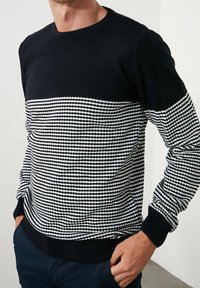 Strickpullover mit einem schwarzen Oberteil und einem weiß-schwarz gestreiften Unterteil. Verfügt über einen Rundhalsausschnitt und gerippte Bündchen.