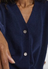 Marinblå stickad cardigan med djup V-hals, prydd med två runda knappar med metallstuk och en mjuk, strukturerad yta.