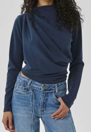 Kobieta w granatowej bluzce z długim rękawem, z marszczoną przednią częścią, w połączeniu z jasnoniebieskimi, wysokim stanem jeansami przewiązanymi cienkim paskiem z denimu.