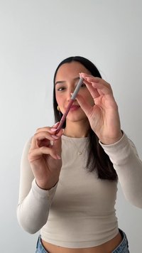Femme aux longs cheveux foncés tient un crayon contour des lèvres mauve près de son visage sur un fond clair uni.