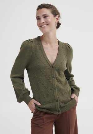 Olivgrüner Strickcardigan mit V-Ausschnitt, strukturiertem Bubbelmuster, Knopfverschluss und eng anliegendem Bündchen, kombiniert mit braunen weit geschnittenen Hosen.