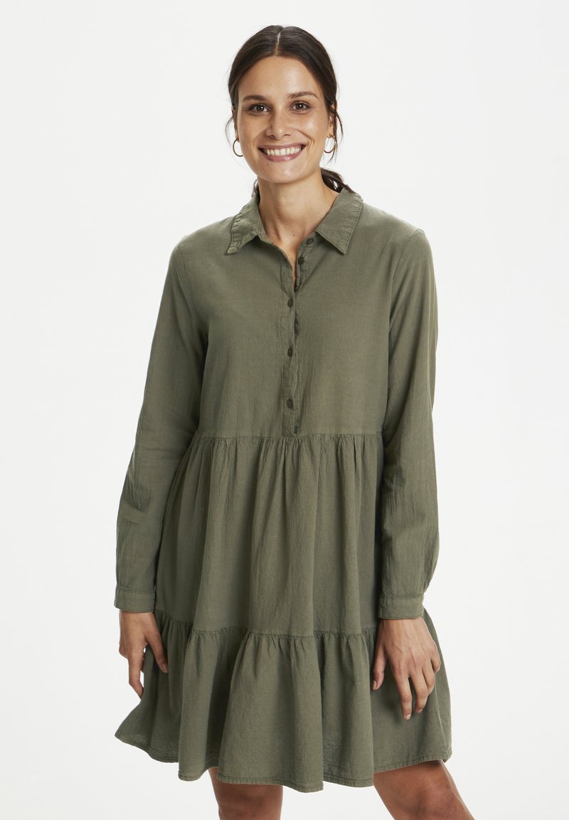 Žena se smiješi, nosi zelenu haljinu dugih rukava s ovratnikom i višeslojnim dijelom suknje, stoji na jednobijeloj pozadini.