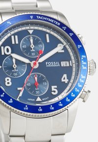 Edelstahl-Chronographenuhr mit blauem Zifferblatt, weißen Ziffern, rotem Sekundenzeiger und Tachymeterlünette. Verfügt über drei Hilfszifferblätter.