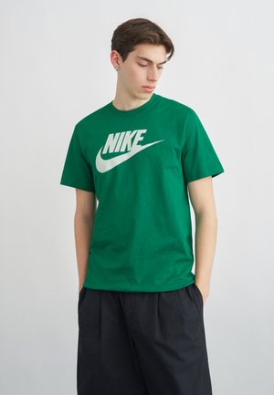 Jeune homme les mains dans les poches portant un t-shirt Nike vert et un pantalon noir ample, regardant vers le bas sur sa gauche sur un fond uni.