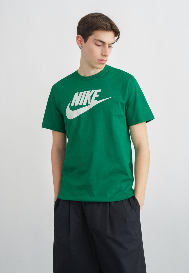 Junger Mann mit den Händen in den Taschen, trägt ein grünes Nike-T-Shirt und lockere schwarze Hosen, blickt nach unten links vor einem einfarbigen Hintergrund.