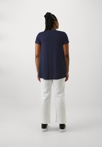 Marineblauw T-shirt met korte mouwen, een ontspannen pasvorm en een ronde zoom, gecombineerd met witte wijde pantalons en zwarte sneakers, gezien vanaf de achterkant.