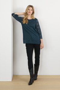 Top in pizzo teal con maniche a tre quarti, orlo ondulato, abbinato a pantaloni neri e stivali da cowboy neri alti, in piedi contro un muro bianco.
