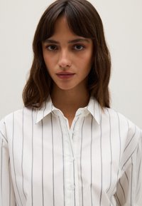Mujer con cabello castaño de longitud media, vistiendo una camisa blanca con finas rayas verticales negras, mirando directamente al frente.