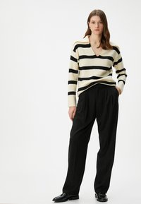Gestreifter Pullover in Creme und Schwarz, mit V-Ausschnitt und gerippten Bündchen, kombiniert mit schwarzen, plissierten Hosen und glänzenden Schuhen.